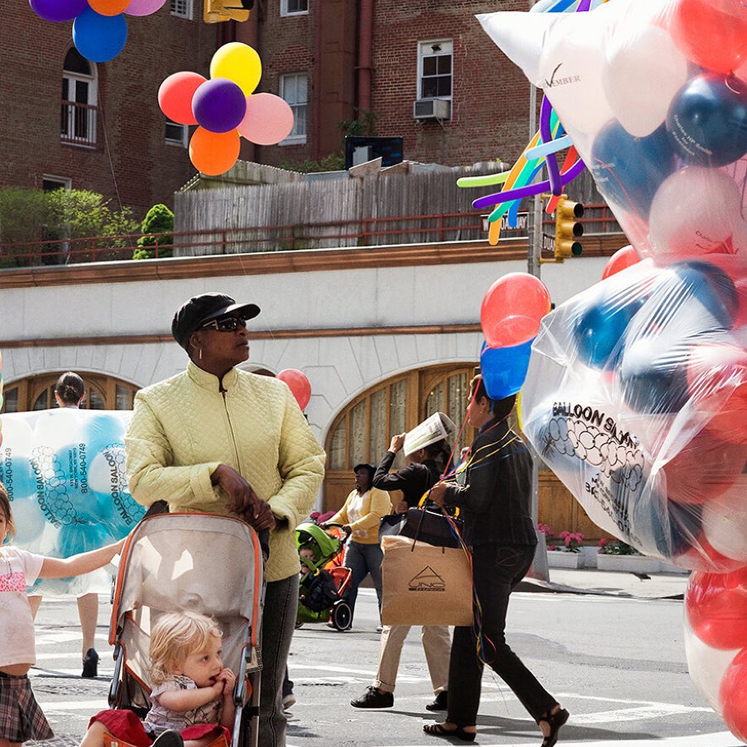 Street scene with an African-American woman pushing a stroller with a young child, accompanied by balloons and other pedestrians.