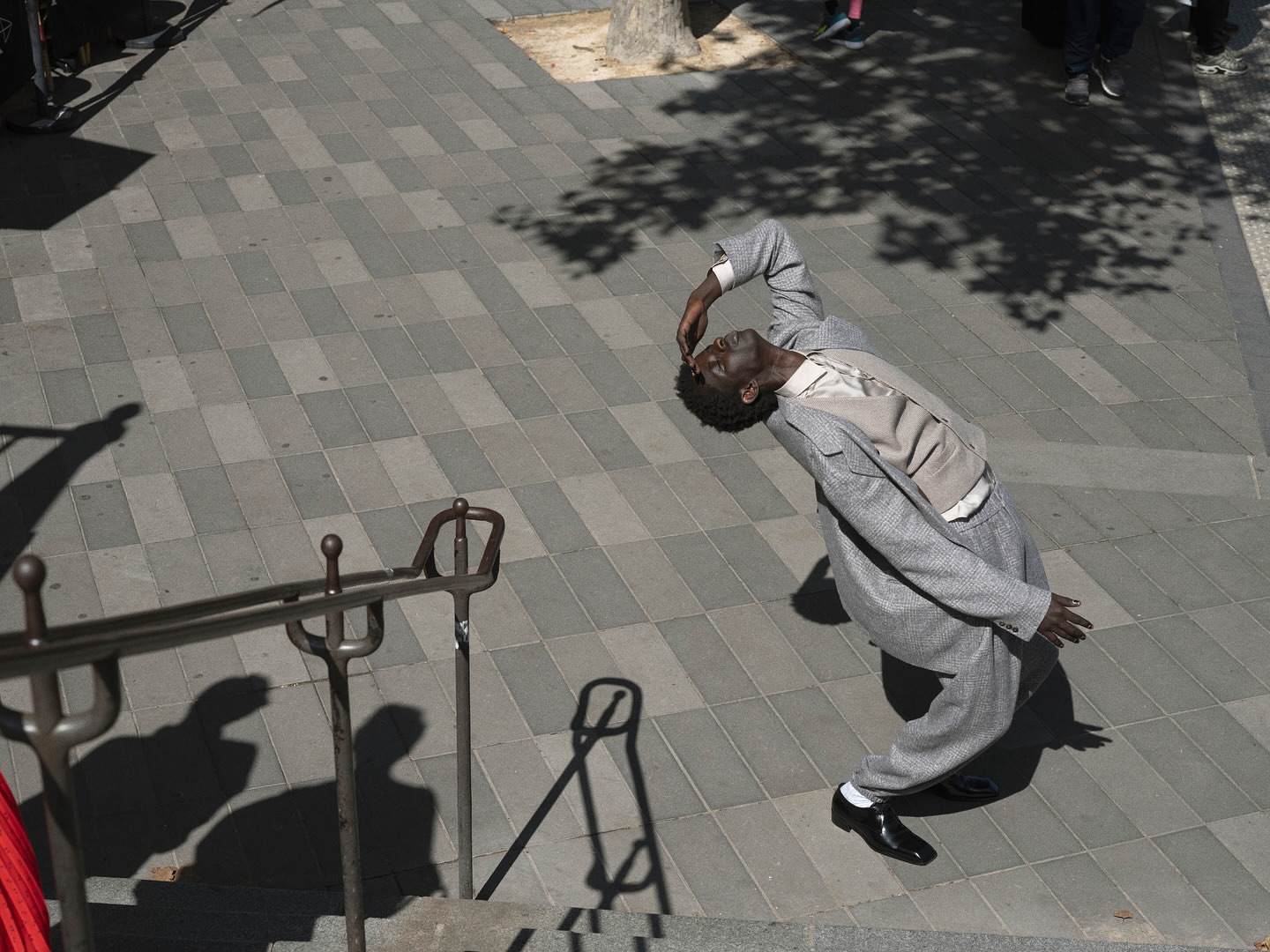 A Black man in a grey suit balances in the sunlight on a city sidewalk while dancing on grey patterned brick near a metal railing.