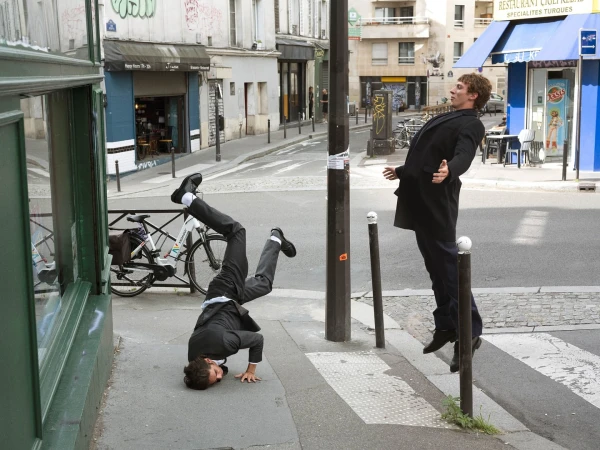 Two men in suits perform stunts on a city street with buildings, storefronts, and a crosswalk in the background on a bright day.