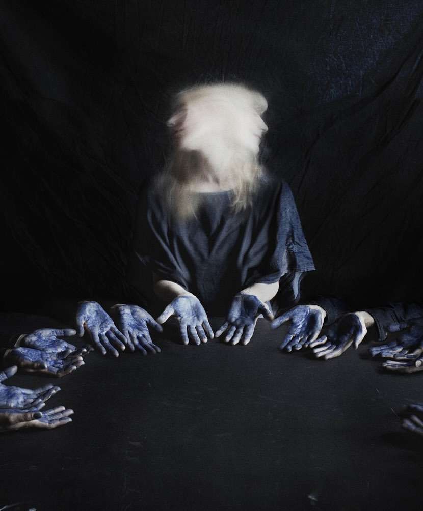 Abstract photo of a person with multiple blurred faces and hands painted blue on a dark table against a shadowy, dark background.