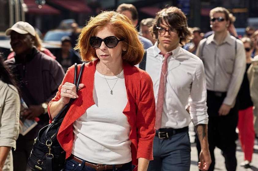 A mature woman with red hair and sunglasses walks alongside a young man in an outdoor urban setting with a crowd of people.