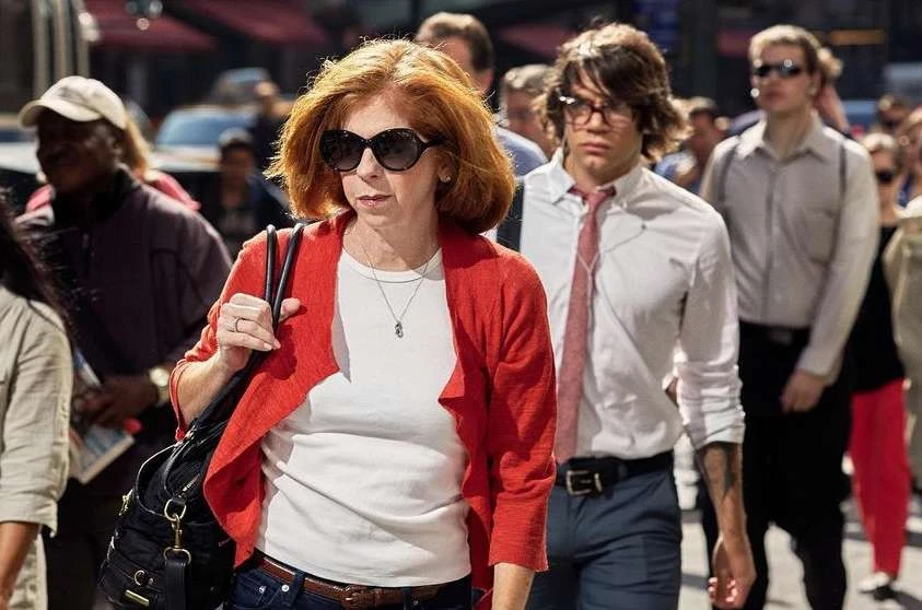 A mature woman with red hair and sunglasses walks alongside a young man in an outdoor urban setting with a crowd of people.