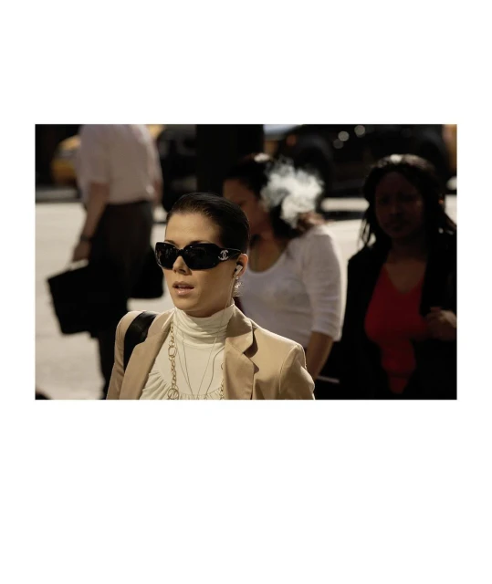 Woman in blazer and sunglasses walks in urban setting with background crowd and cars, neutral color palette, medium shot.