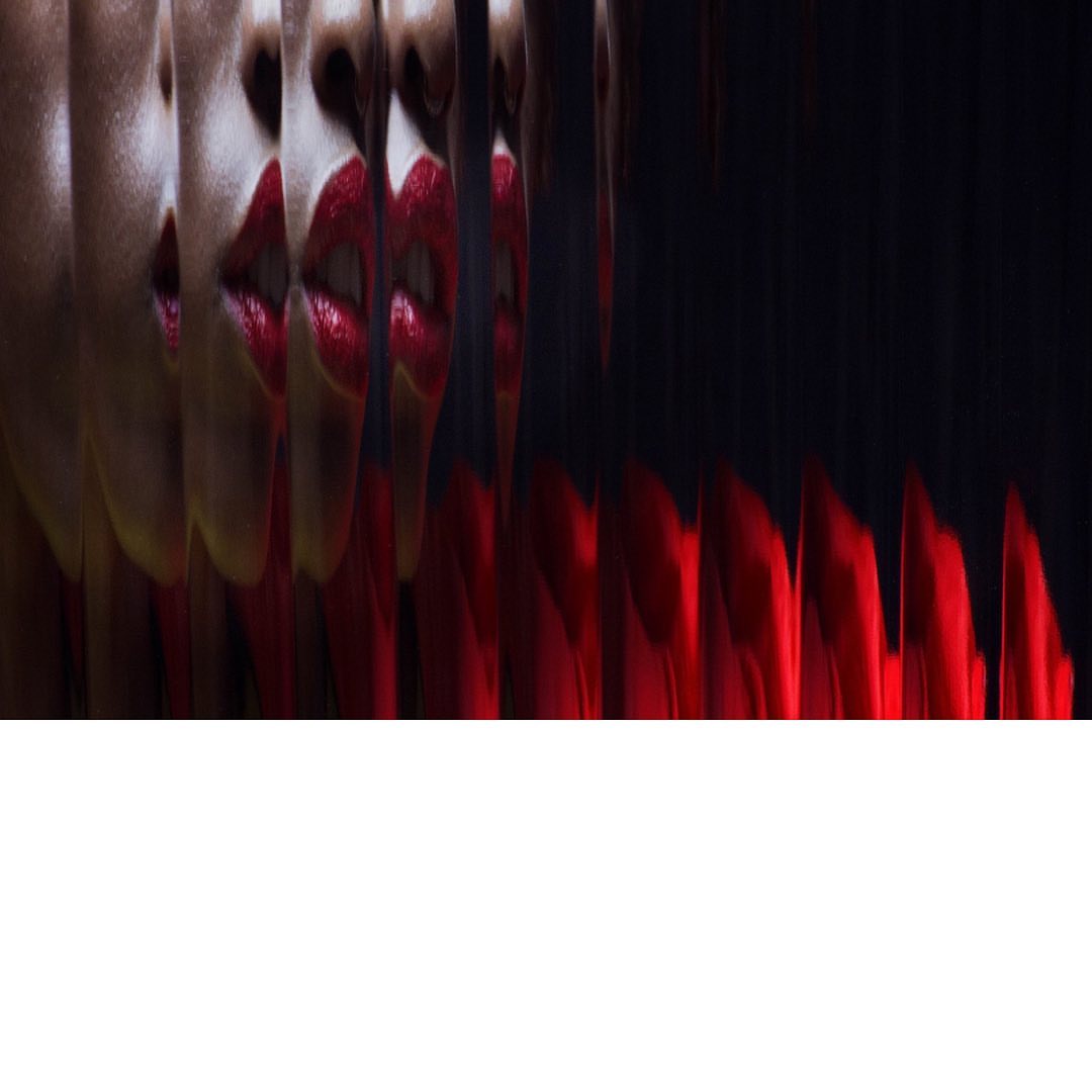 Extreme close-up of a woman's face with bright red lipstick, distorted into vertical bands against a dark, abstract background.
