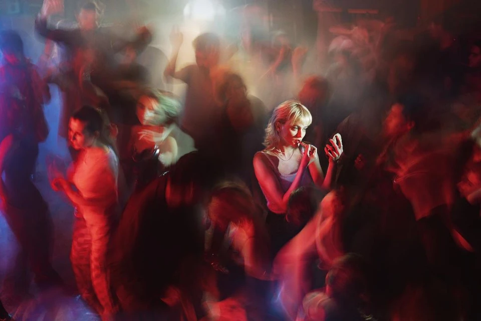 Woman applies lipstick in a crowded nightclub, people dance in the background, illuminated by red and blue lights, creating a vibrant atmosphere.