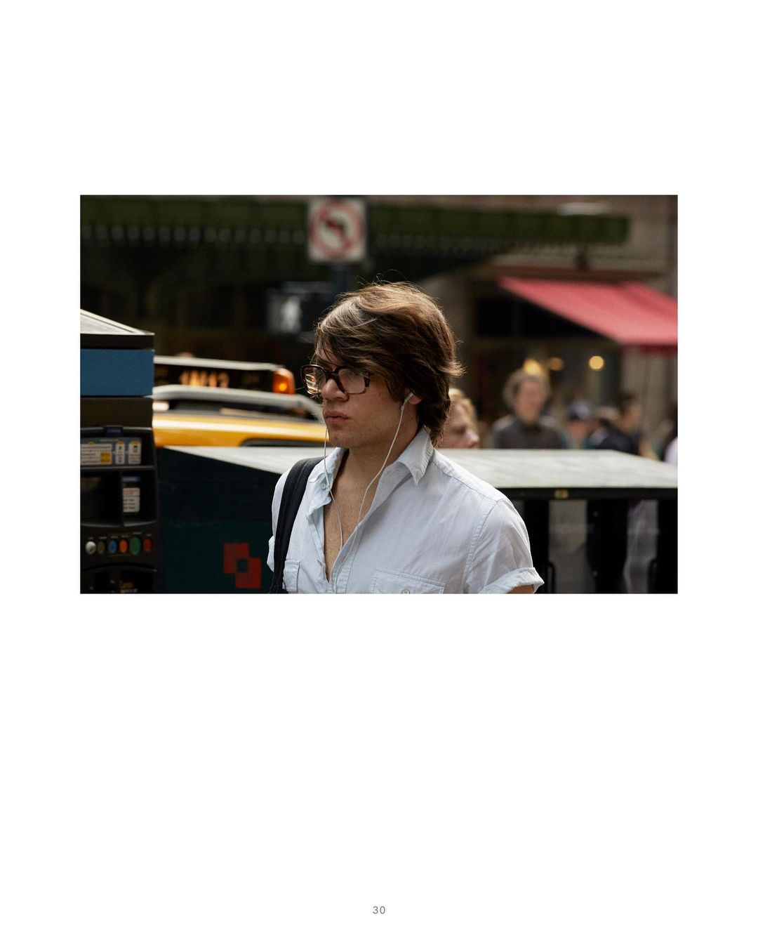 Young man with dark hair wears glasses and ear buds in an urban setting with a yellow taxi and building in soft-focus background.