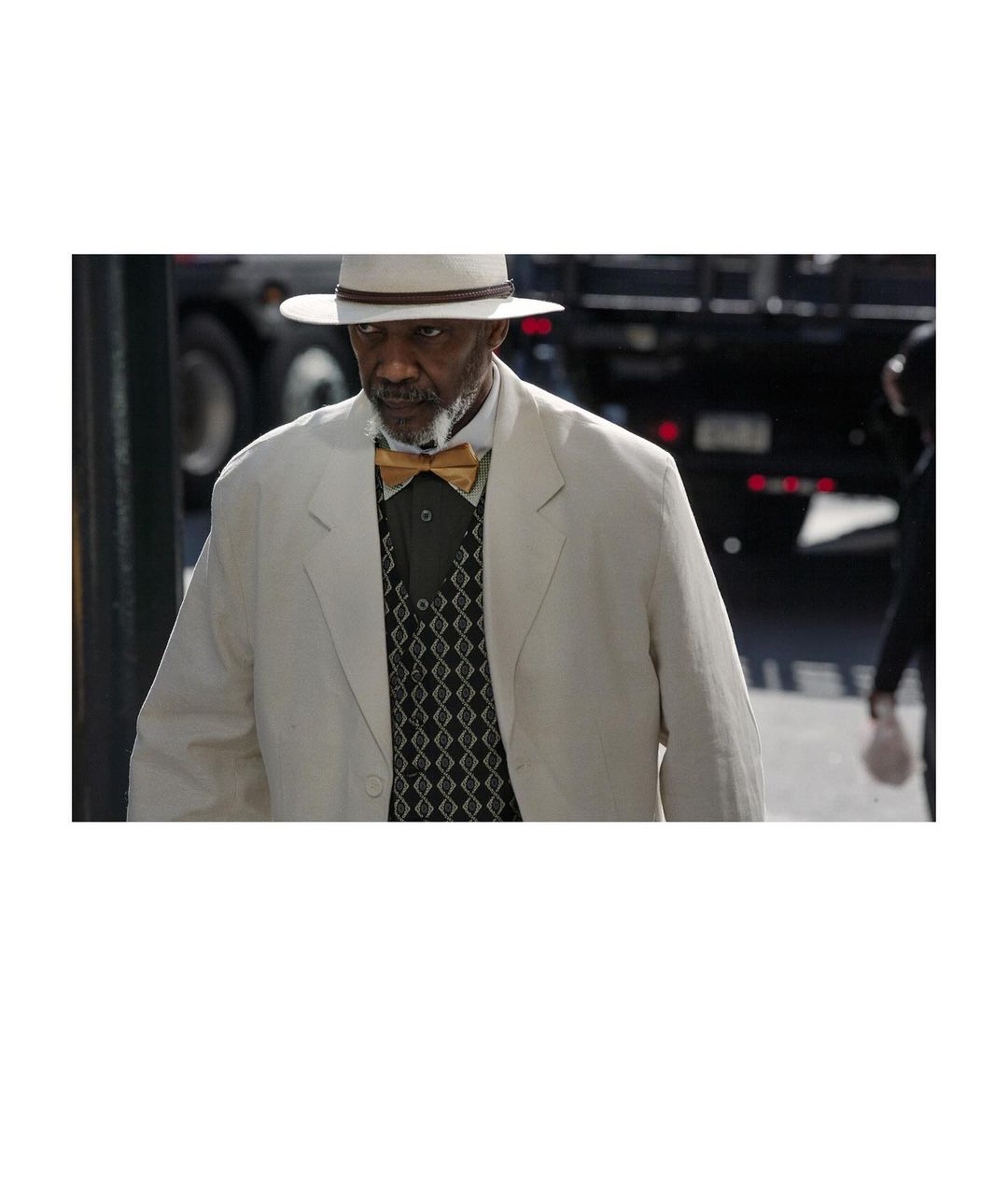 Mature Black man in a cream suit and Panama hat poses outdoors, wearing a gold bow tie and patterned vest, with a truck in the background.
