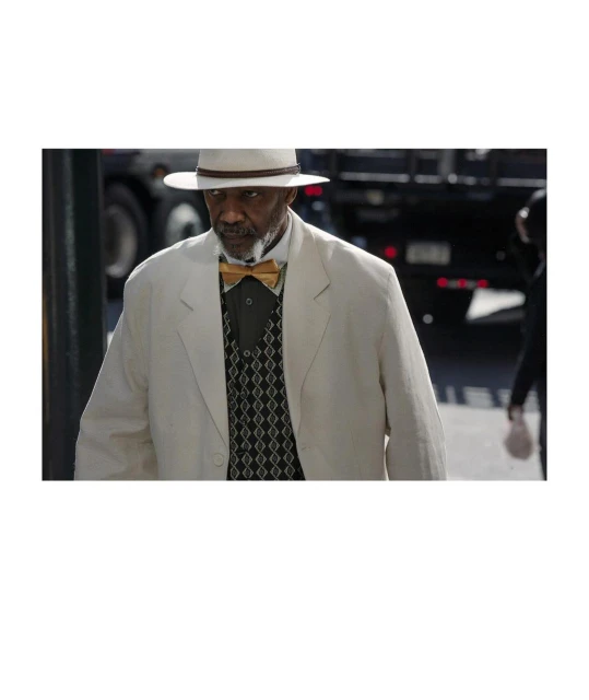Mature Black man in a cream suit and Panama hat poses outdoors, wearing a gold bow tie and patterned vest, with a truck in the background.