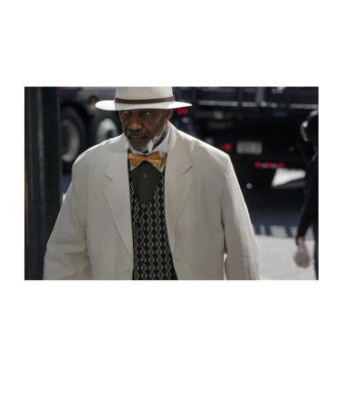 Mature Black man in a cream suit and Panama hat poses outdoors, wearing a gold bow tie and patterned vest, with a truck in the background.