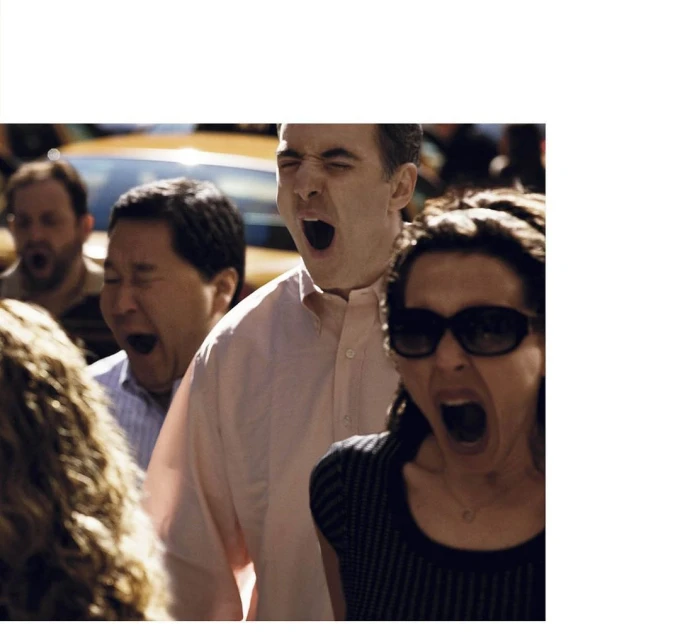 Diverse group of people with open mouths, seemingly yawning or shouting, suggesting a crowd scene, perhaps in a city setting during daylight.
