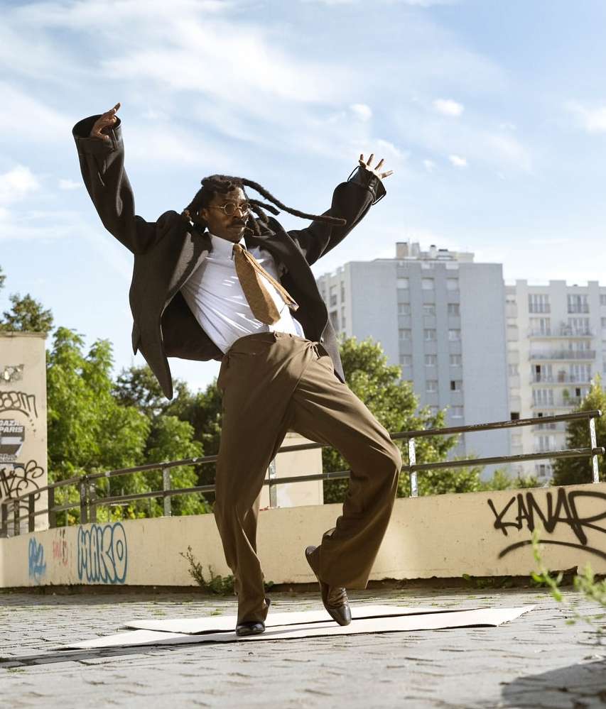Adult male with dreadlocks in suit dancing on concrete surface in urban setting with blue sky, buildings, and greenery visible.