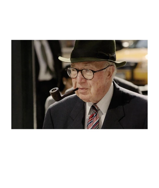 Elderly man with hat and glasses smoking a pipe, wearing a dark suit and patterned tie, against a blurred background.