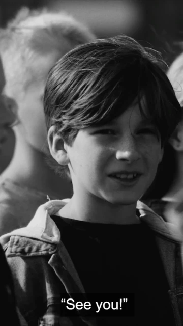 Black and white portrait of a child with medium brown hair and a denim jacket, looking at the camera, with other kids in background.