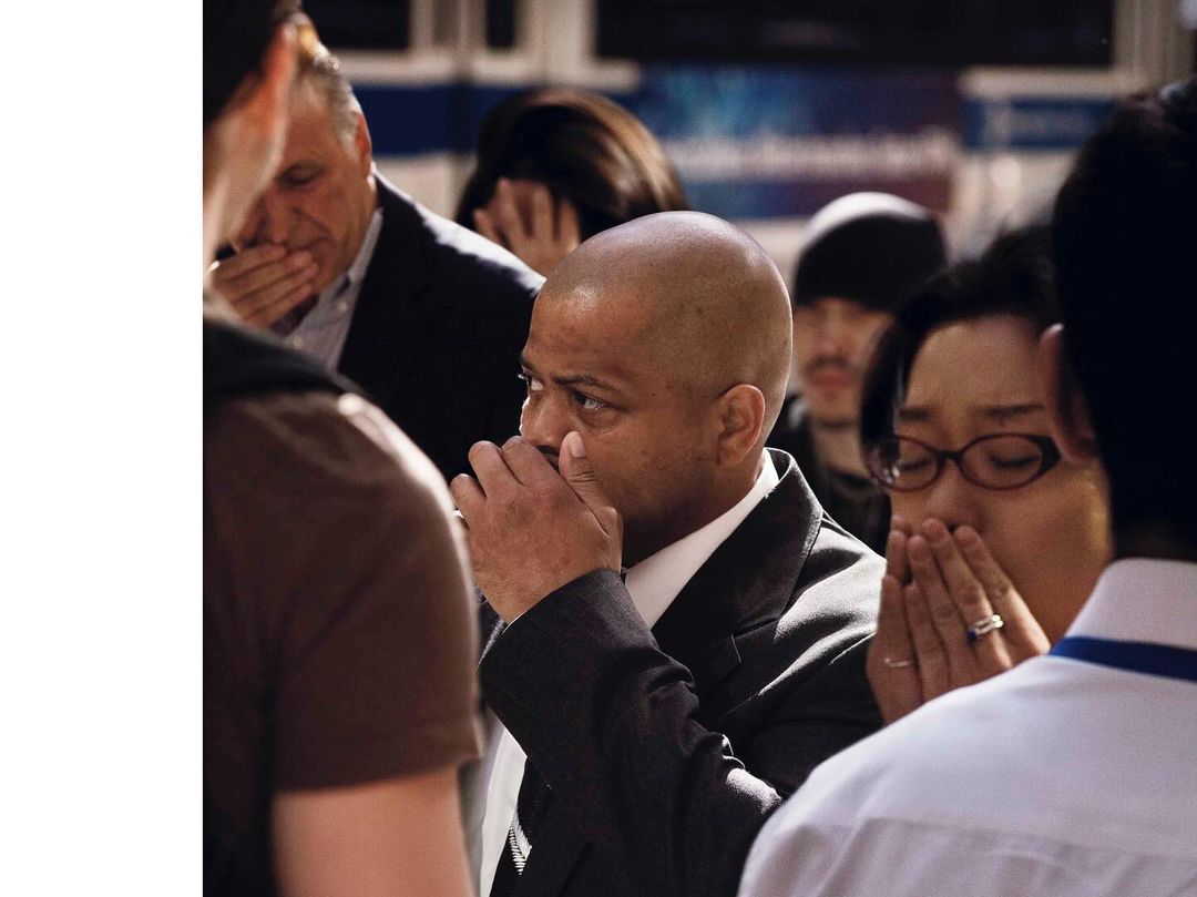 Candid photo of a diverse group of people in suits and dresses, covering their faces with their hands, in a brightly-lit setting.