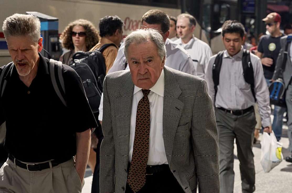 A medium shot of an adult male wearing a patterned blazer and tie walking outside, surrounded by a crowd of people in an urban setting.