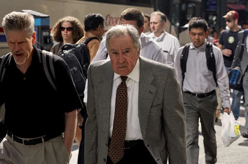 A medium shot of an adult male wearing a patterned blazer and tie walking outside, surrounded by a crowd of people in an urban setting.