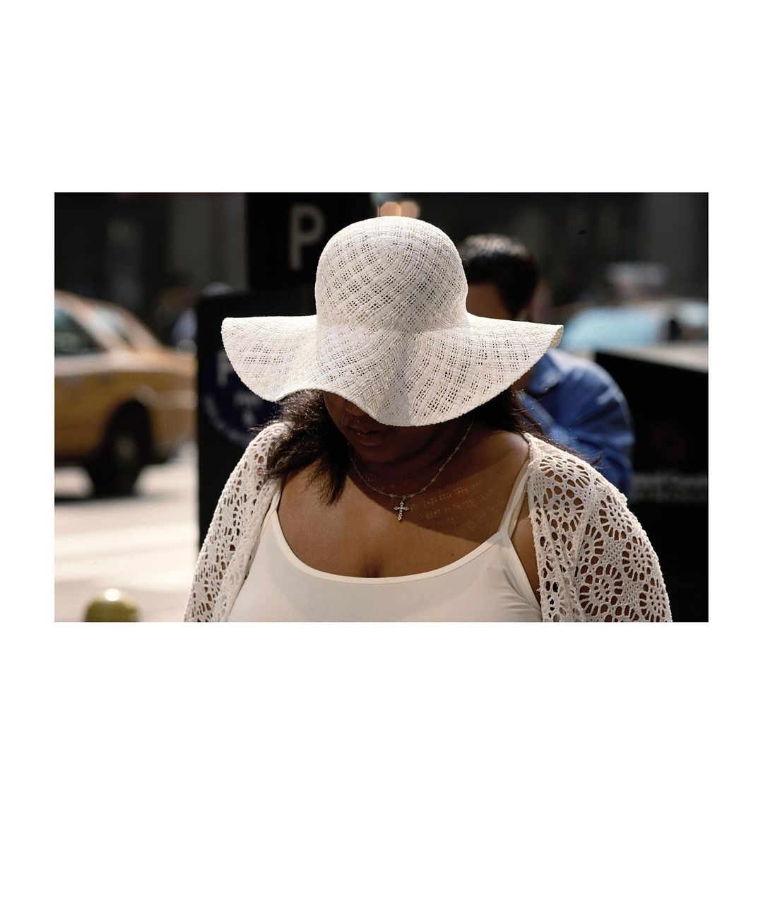 Woman in white sun hat and lace cardigan stands in city street with yellow taxi and blurry pedestrians in background on sunny day.