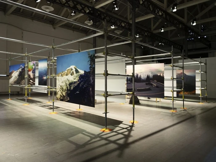An art exhibition installation inside a large industrial warehouse featuring large photographs mounted on scaffolding structures.