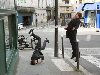 Two men in dark suits on a city sidewalk, one headspinning on the ground and one floating upright.