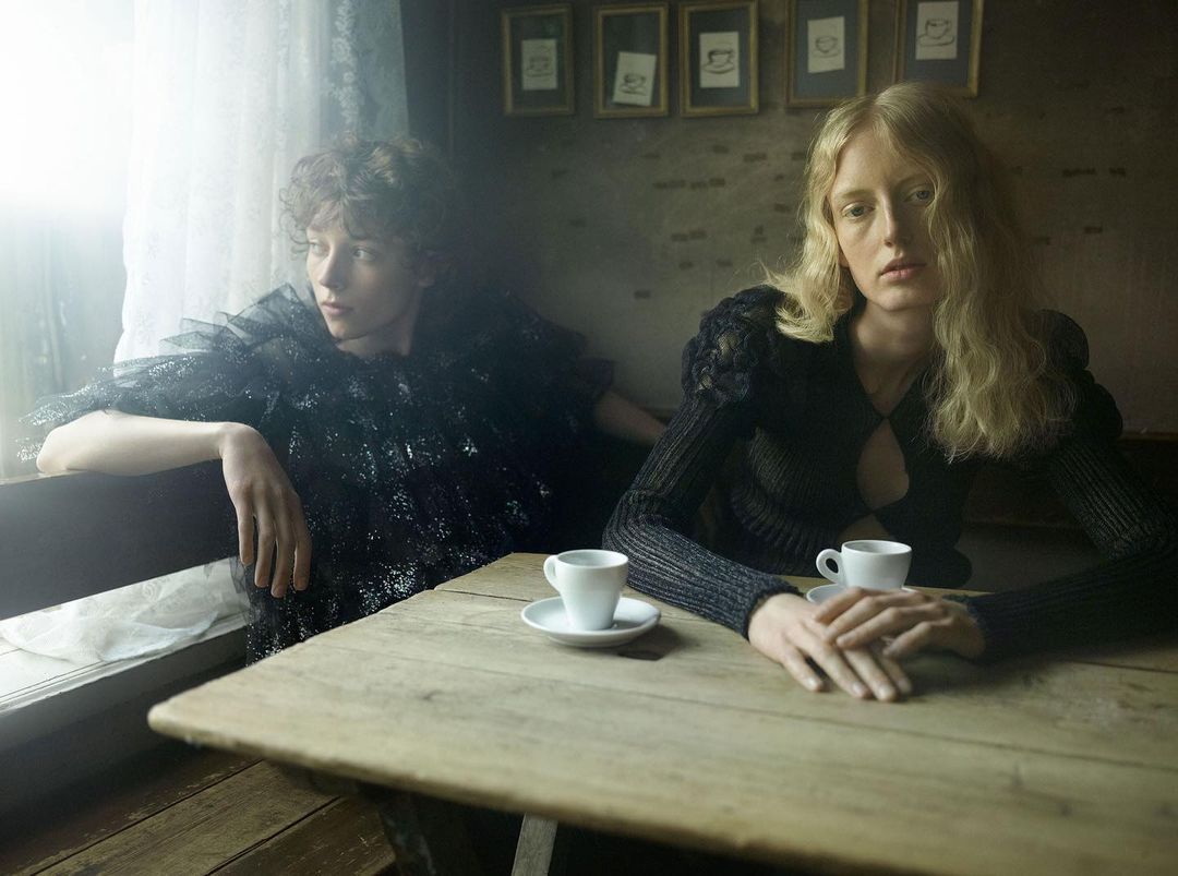 Two women in dark fashionable clothing sit at a wooden table in a cafe, with white espresso cups and a muted, intimate atmosphere.