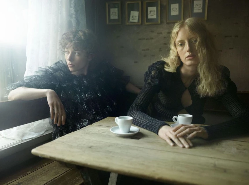 Two women in dark fashionable clothing sit at a wooden table in a cafe, with white espresso cups and a muted, intimate atmosphere.