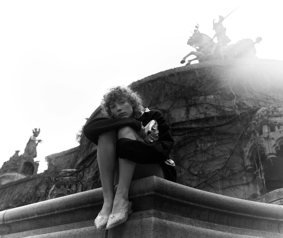 Young woman sits on a ledge with sculpture in background in a black and white outdoor fashion portrait with neutral facial expression.