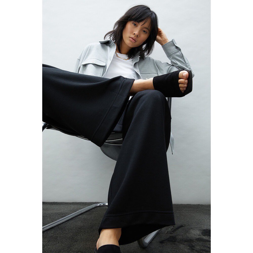 Fashion portrait of an Asian woman wearing a light blue jacket and black wide-leg pants sitting on a chair against a white wall.