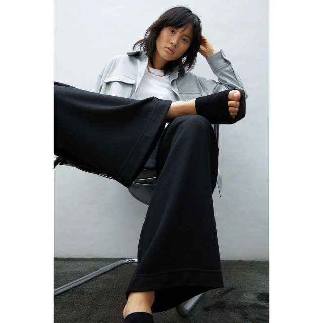 Fashion portrait of an Asian woman wearing a light blue jacket and black wide-leg pants sitting on a chair against a white wall.