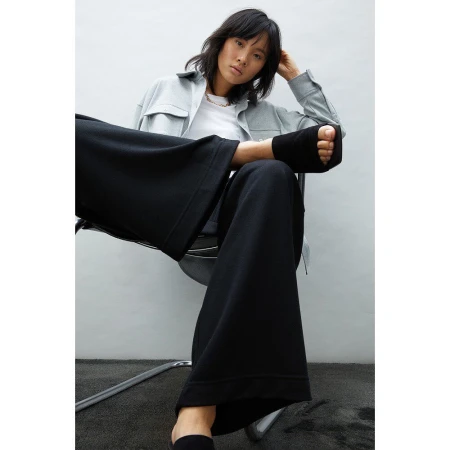 Fashion portrait of an Asian woman wearing a light blue jacket and black wide-leg pants sitting on a chair against a white wall.