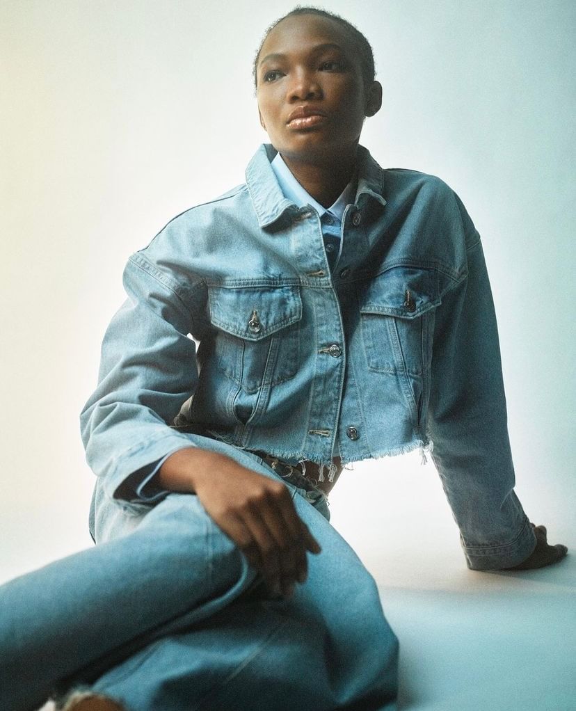 Black woman with short hair wearing a denim jacket and jeans seated against a pale backdrop