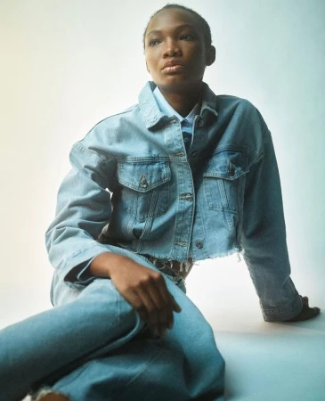 Black woman with short hair wearing a denim jacket and jeans seated against a pale backdrop