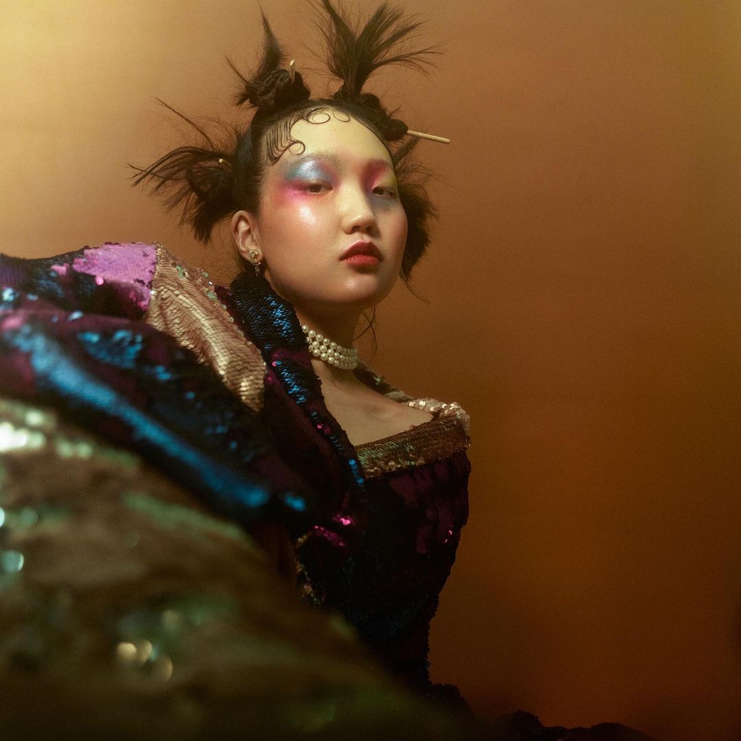 Portrait of an Asian female model with creative makeup, wearing a sequined garment and pearl necklace against a gold-toned background.