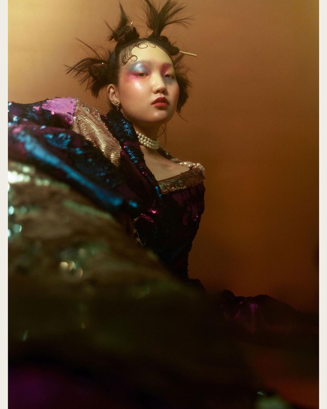 Eye-level portrait of an Asian female wearing a sequined top and pearls against an orange background in a fashion photography setting.