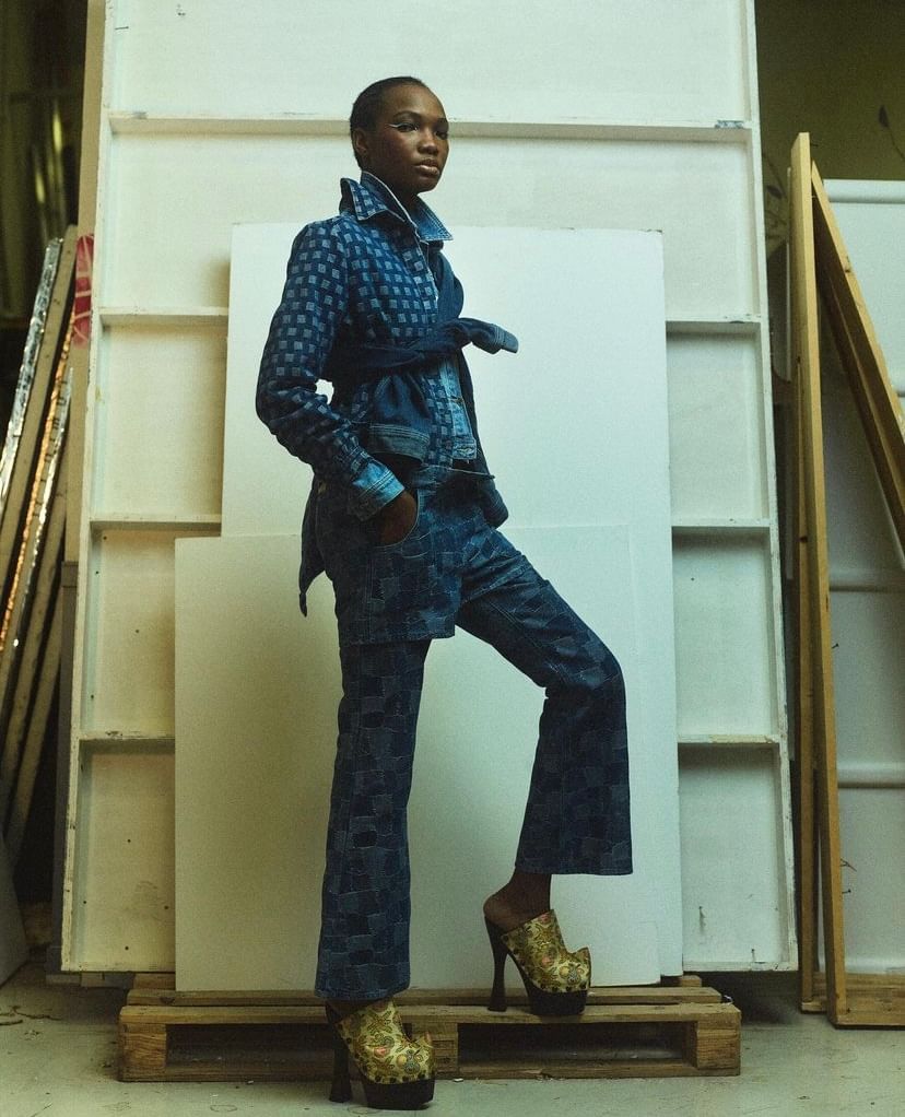 Fashion model in patterned denim jacket and pants posing in a photo studio with a backdrop on wooden pallets, wearing platform heels.