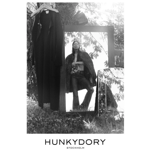 An Asian woman in jeans and a coat stands in a grassy outdoor setting, reflected in mirrors alongside a hanging black coat, styled in black and white.