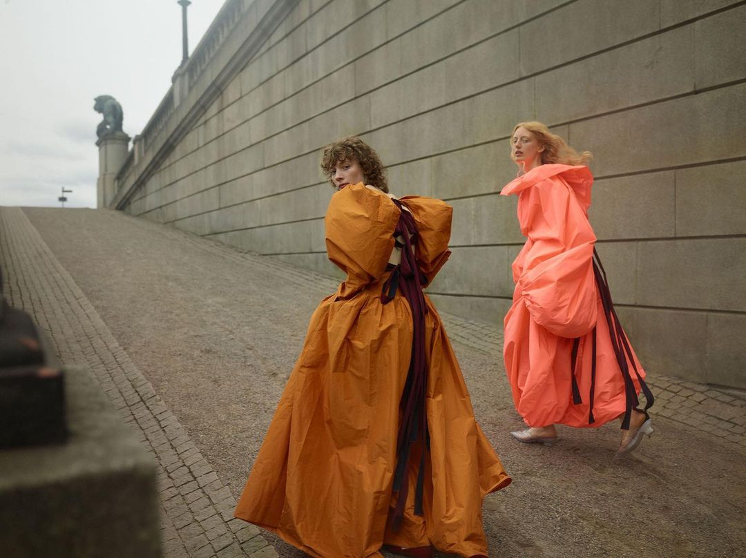Two women in orange and peach balloon gowns walk along a stone path in a muted outdoor setting. Fashion-forward outdoor editorial.