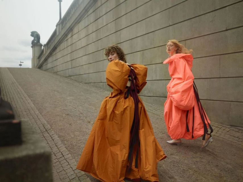 Two women in orange and peach balloon gowns walk along a stone path in a muted outdoor setting. Fashion-forward outdoor editorial.