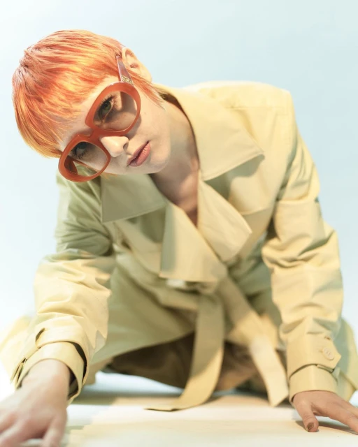 Stylish young woman with red hair and oversized red glasses poses in a light tan trench coat against a pale background in a fashion studio.