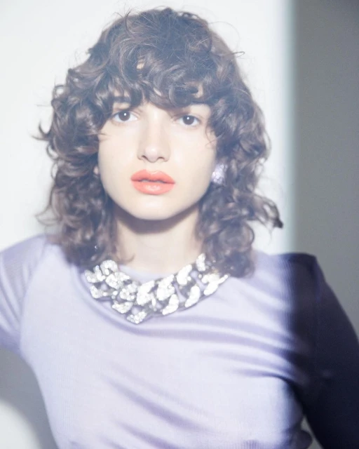 Portrait of a young woman with curly hair wearing a light purple sweater and a chunky silver necklace against a light background.