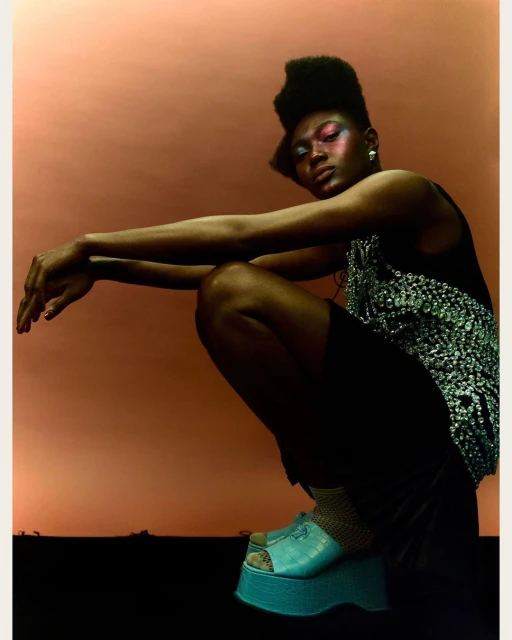 Woman with colorful eye makeup and platform sandals strikes a low-angle pose against a peach background in a fashion editorial shot.