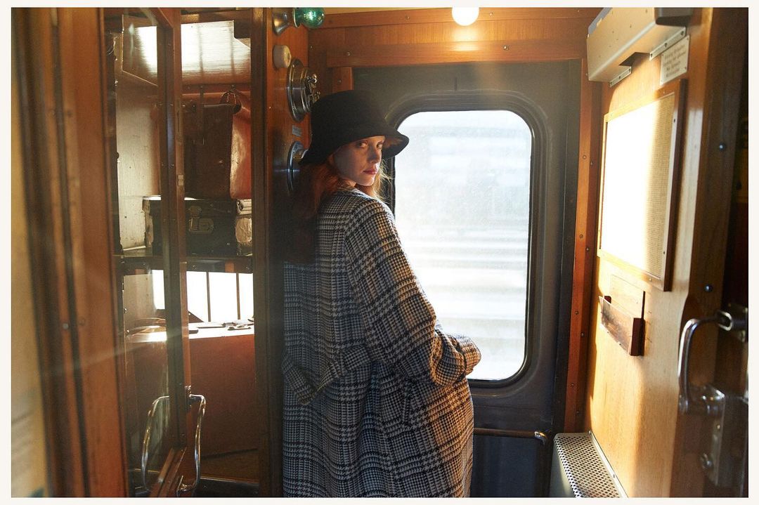 Woman in a plaid coat and black bucket hat standing inside a wood-paneled train compartment looking back