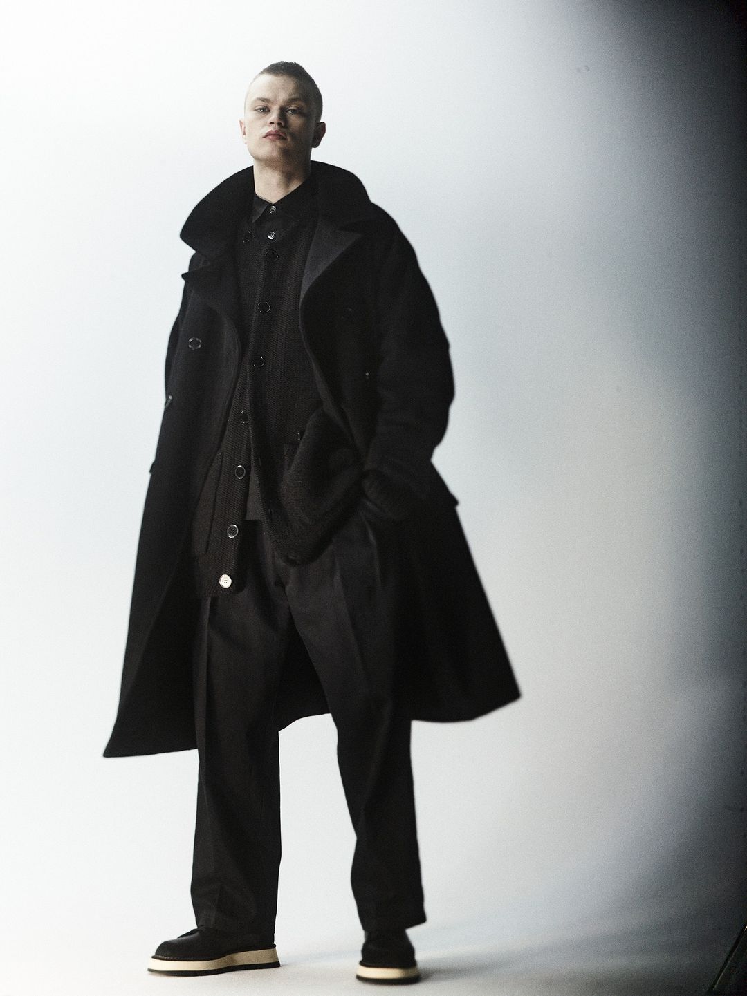 Adult male fashion model posing indoors wearing a black coat, shirt, and pants, styled with dark shoes against a gradient background.