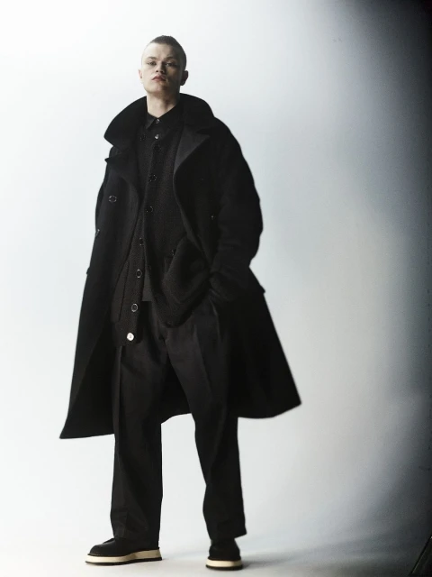 Adult male fashion model posing indoors wearing a black coat, shirt, and pants, styled with dark shoes against a gradient background.