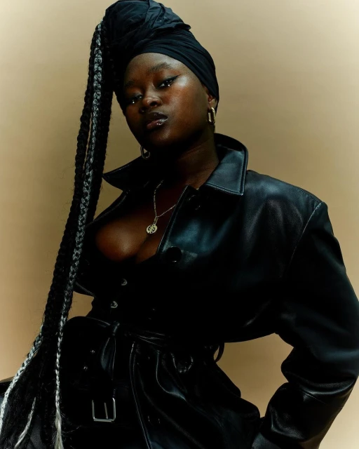 Stylish portrait of a woman with dark skin and long braids, wearing a black headwrap and leather jacket against a neutral backdrop.