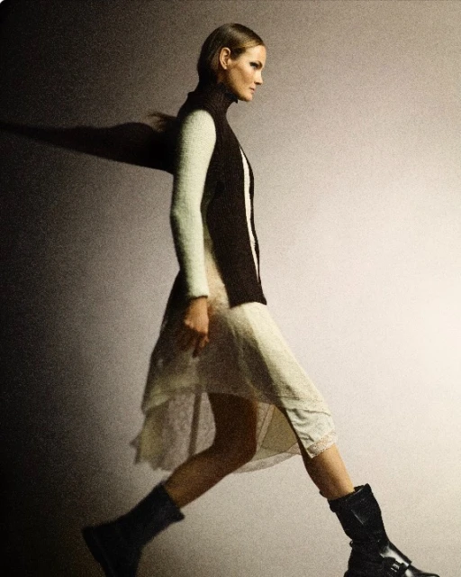 Fashion editorial of a woman walking and wearing a sweater vest, dress, and boots in a photo studio with dramatic lighting.