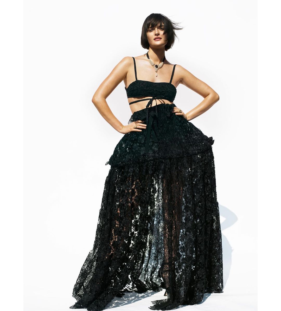 A woman in a black lace crop top and full-length skirt poses against a bright background, showcasing a high-fashion editorial look.