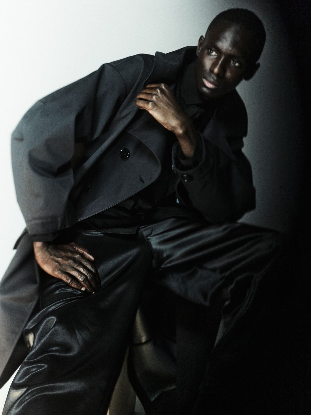 Dark-skinned male model in a black trench coat and shiny black pants poses in a photo studio against a graduated white-to-black background.