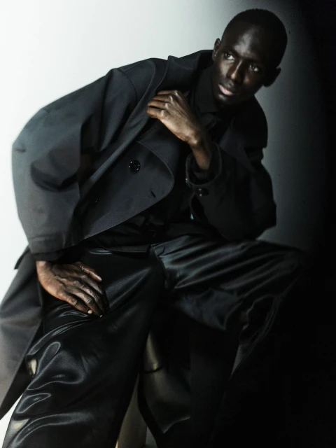 Dark-skinned male model in a black trench coat and shiny black pants poses in a photo studio against a graduated white-to-black background.