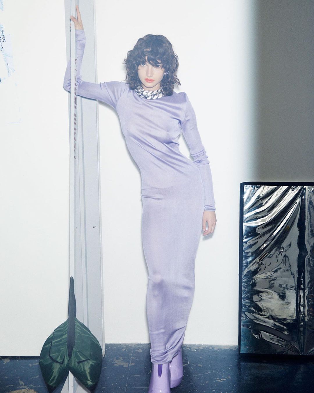 A female fashion model with curly hair poses in a long-sleeved lavender dress and matching lavender boots in a studio setting.