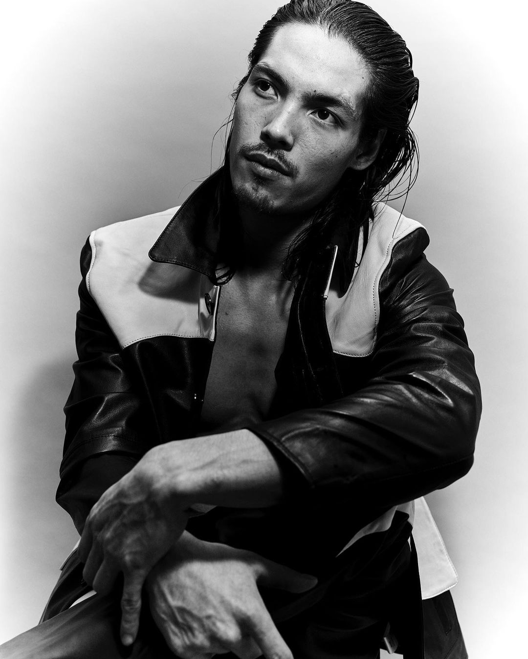 Black and white portrait of a man with long dark hair, wearing a leather jacket with white panels, sitting indoors with a neutral expression.
